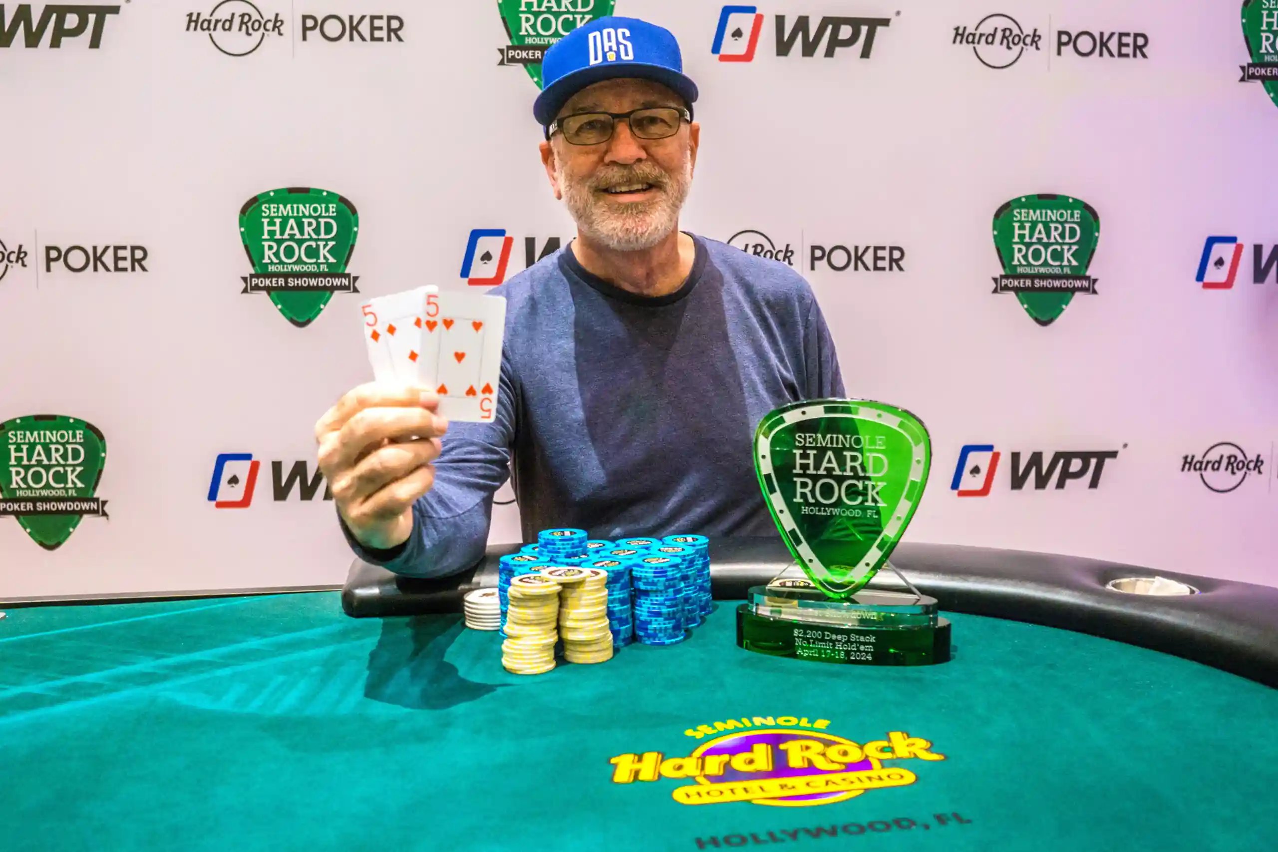 Man seated at a poker table holds up pocket fives beside stacks of chips and a green Seminole Hard Rock Poker Showdown trophy, with WPT and Hard Rock Poker logos on the backdrop.
