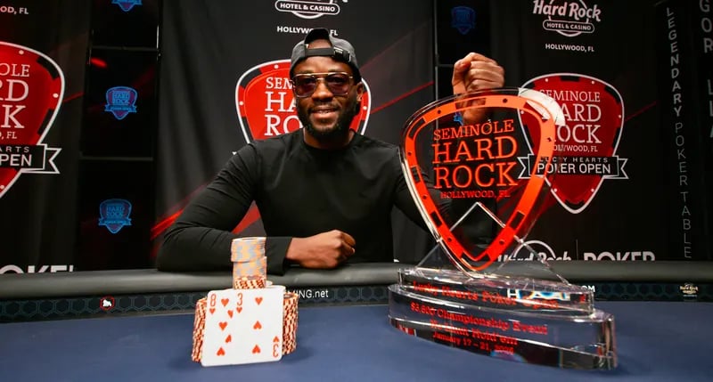 Smiling poker champion holds Seminole Hard Rock Lucky Hearts Poker Open trophy beside winning hand and chips.