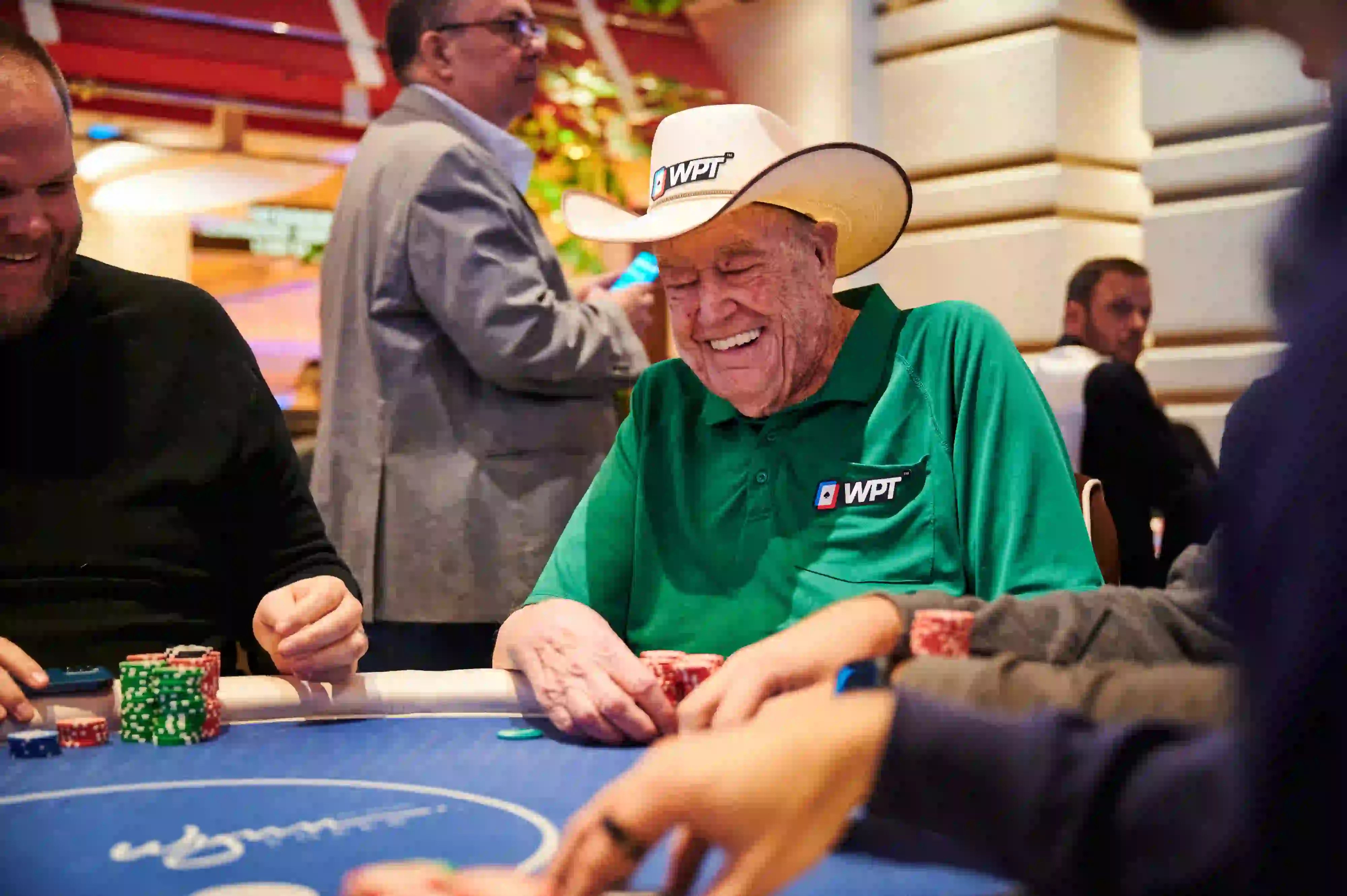 An older man wearing a cowboy hat and a green WPT shirt laughs while playing poker at a casino table, surrounded by other players and stacks of chips.