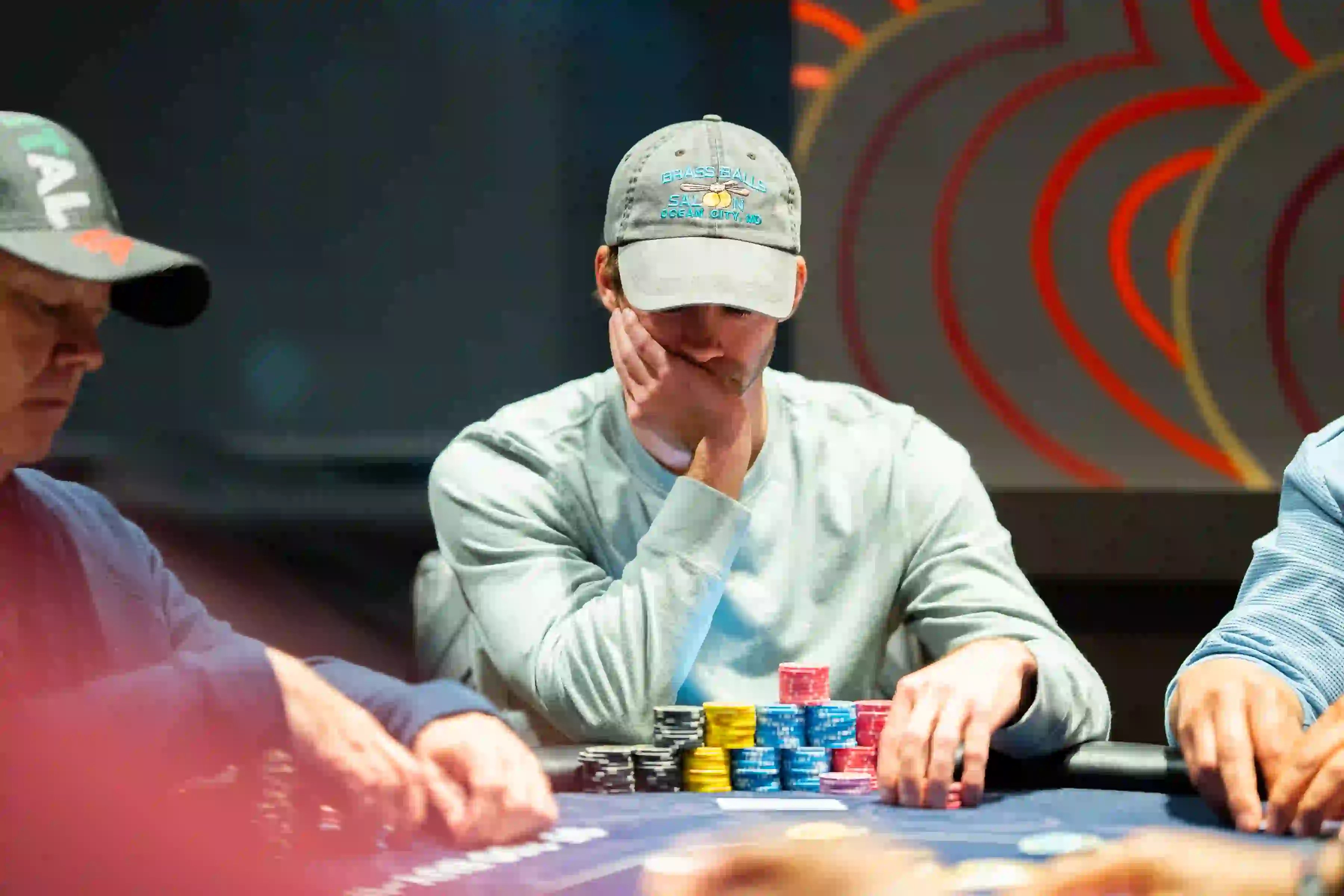 A poker player in a light grey cap and long-sleeve shirt sits at a table with a large stack of multicolored chips, thinking through a hand during live tournament play.