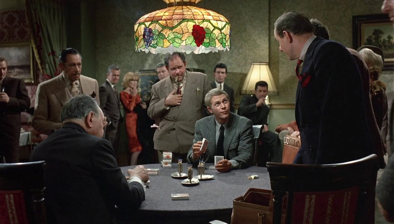Players gather around a vintage poker table as Steve McQueen’s character studies his cards in a crowded room in The Cincinnati Kid.