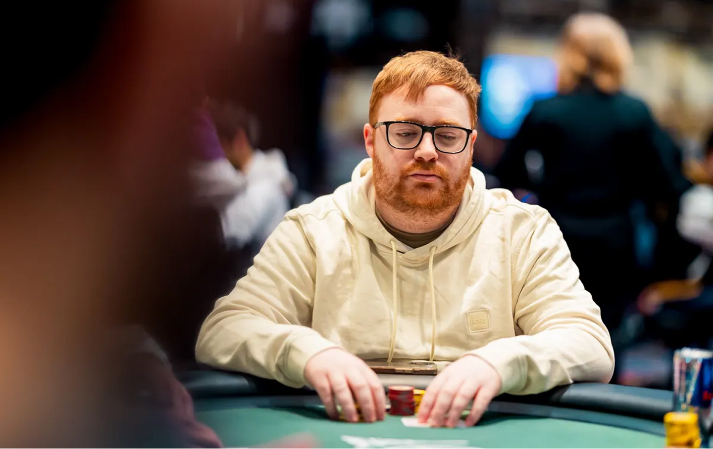 Killian Farrell in a light hoodie at a tournament table, focused on gameplay with blurred players in the background.