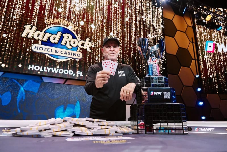 WPT® champion at Seminole Hard Rock Hollywood holding winning hand beside trophy and cash bricks on final table.
