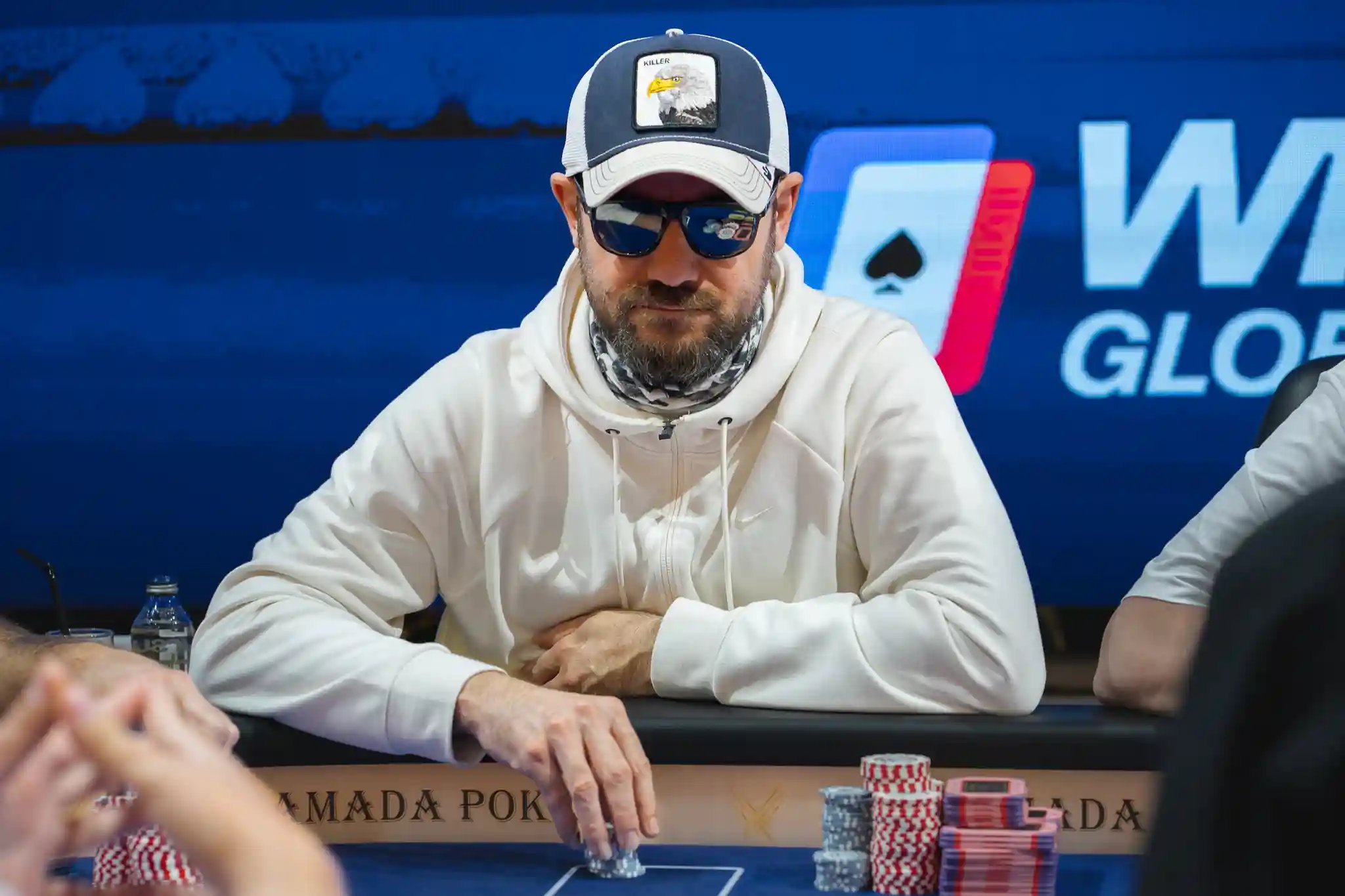 Poker player sitting at the table in a white hoodie and cap, wearing sunglasses, stacking chips during a WPT® Global event.