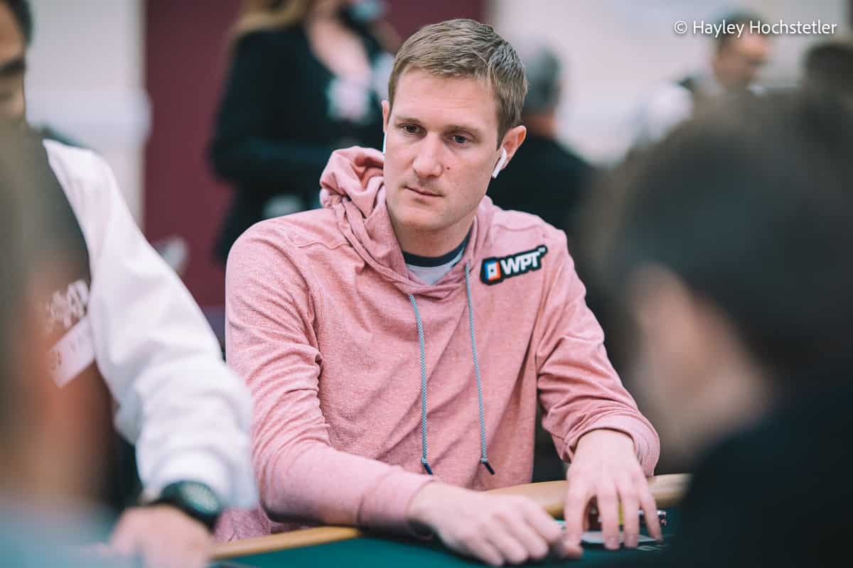 Brad Owen wearing a pink WPT hoodie sits at a tournament table, focused on the hand in front of him. Other players and a dealer are blurred in the background.