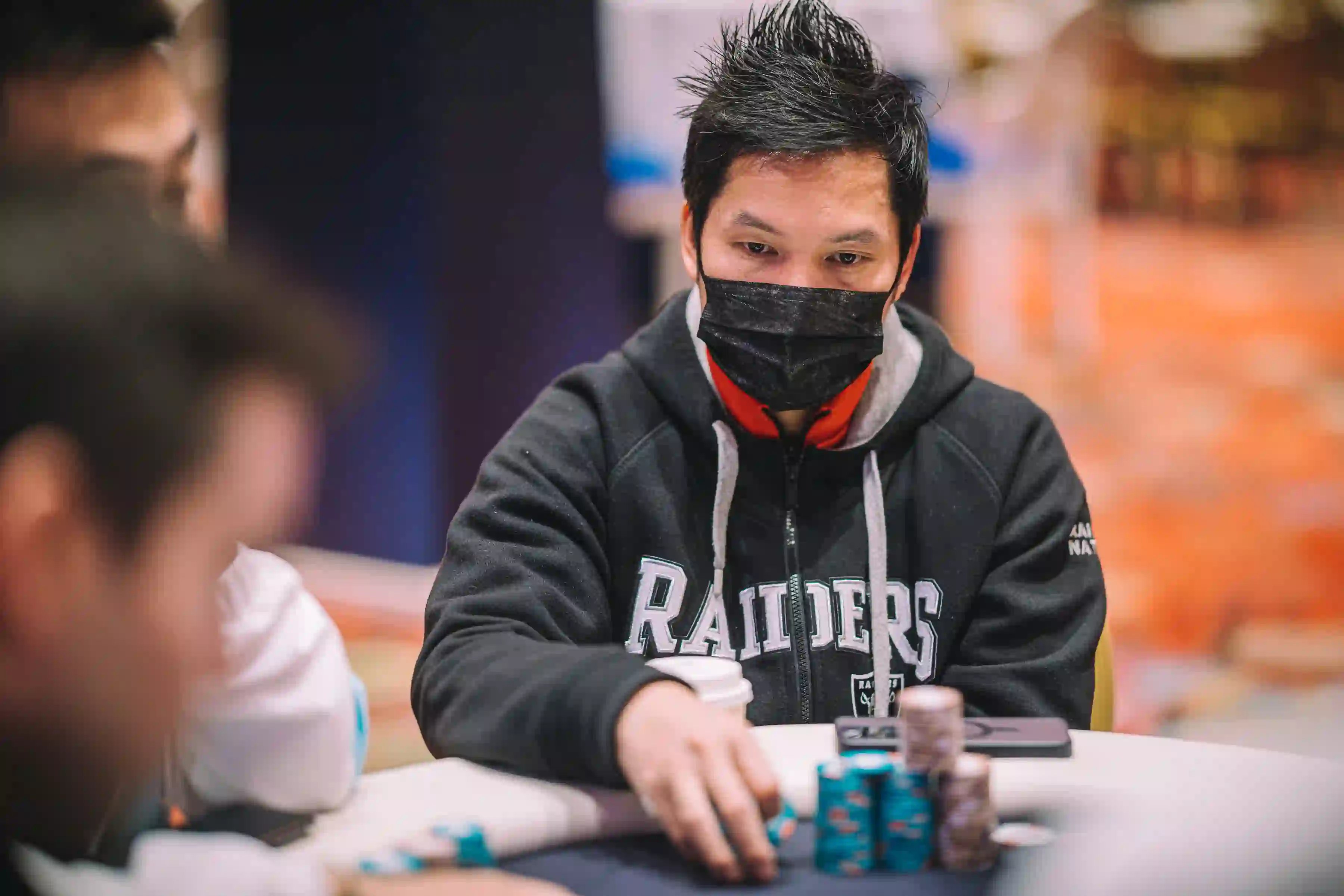 Focused poker player in Raiders hoodie and face mask stacking chips during live tournament.