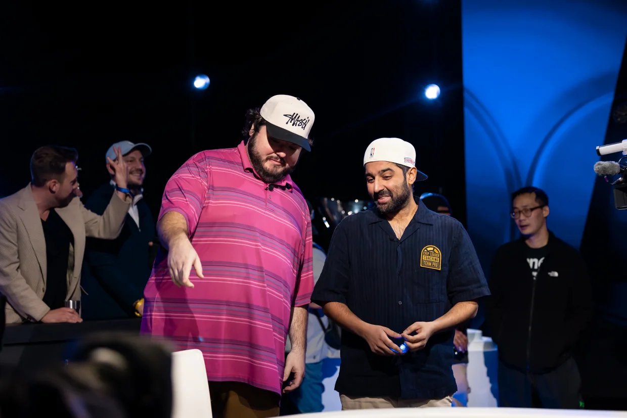 Two friends in casual caps stand by WPT® feature table, discussing action with rail behind.
