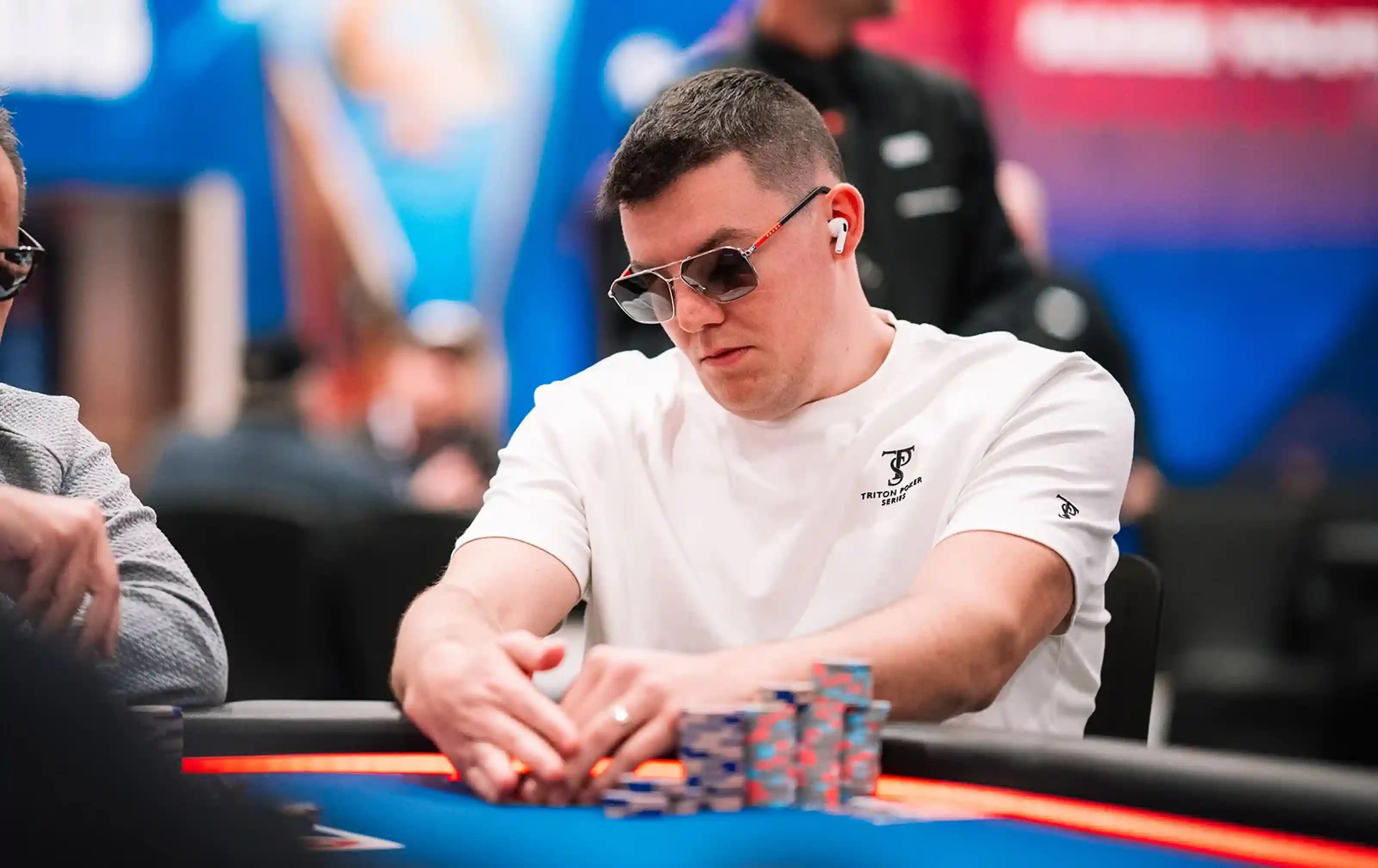 Jesse Lonis stacking chips during a major poker tournament, wearing sunglasses and a white Triton Poker Series shirt.