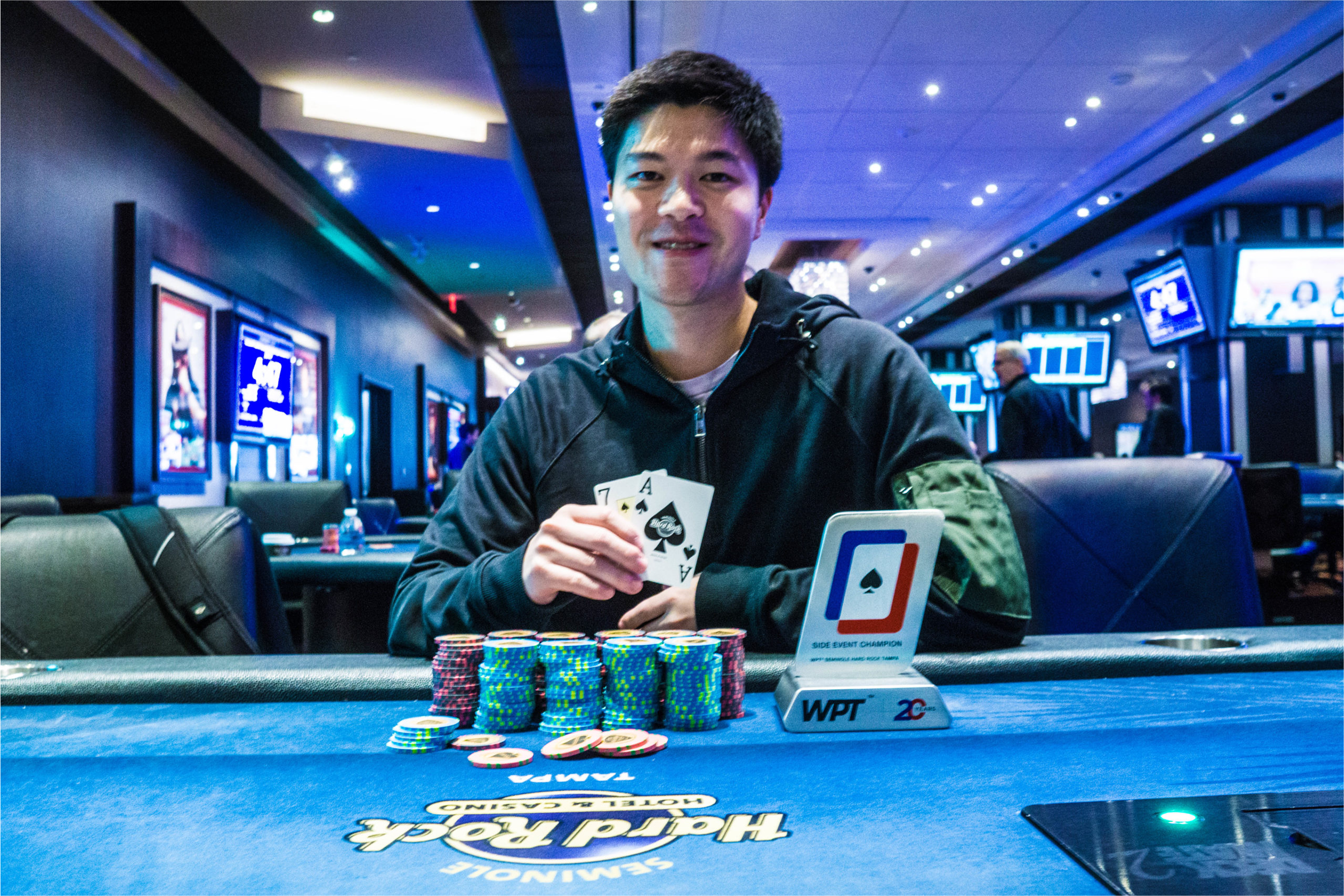 Clemen Deng smiling while holding cards and posing with WPT trophy and chips on the table.