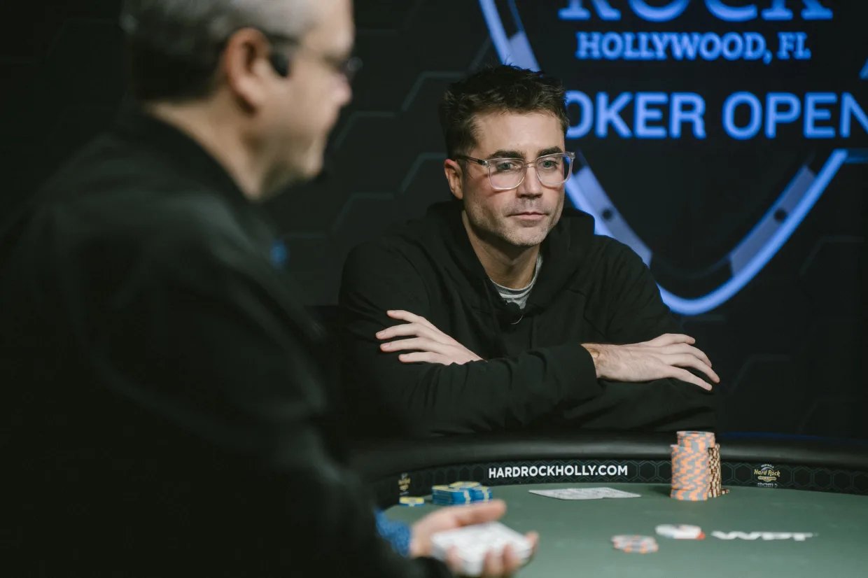 Kevin Nee sits at the final table of the Seminole Hard Rock Poker Open, arms crossed and focused, with tournament chips in front of him and the event logo glowing in the background.