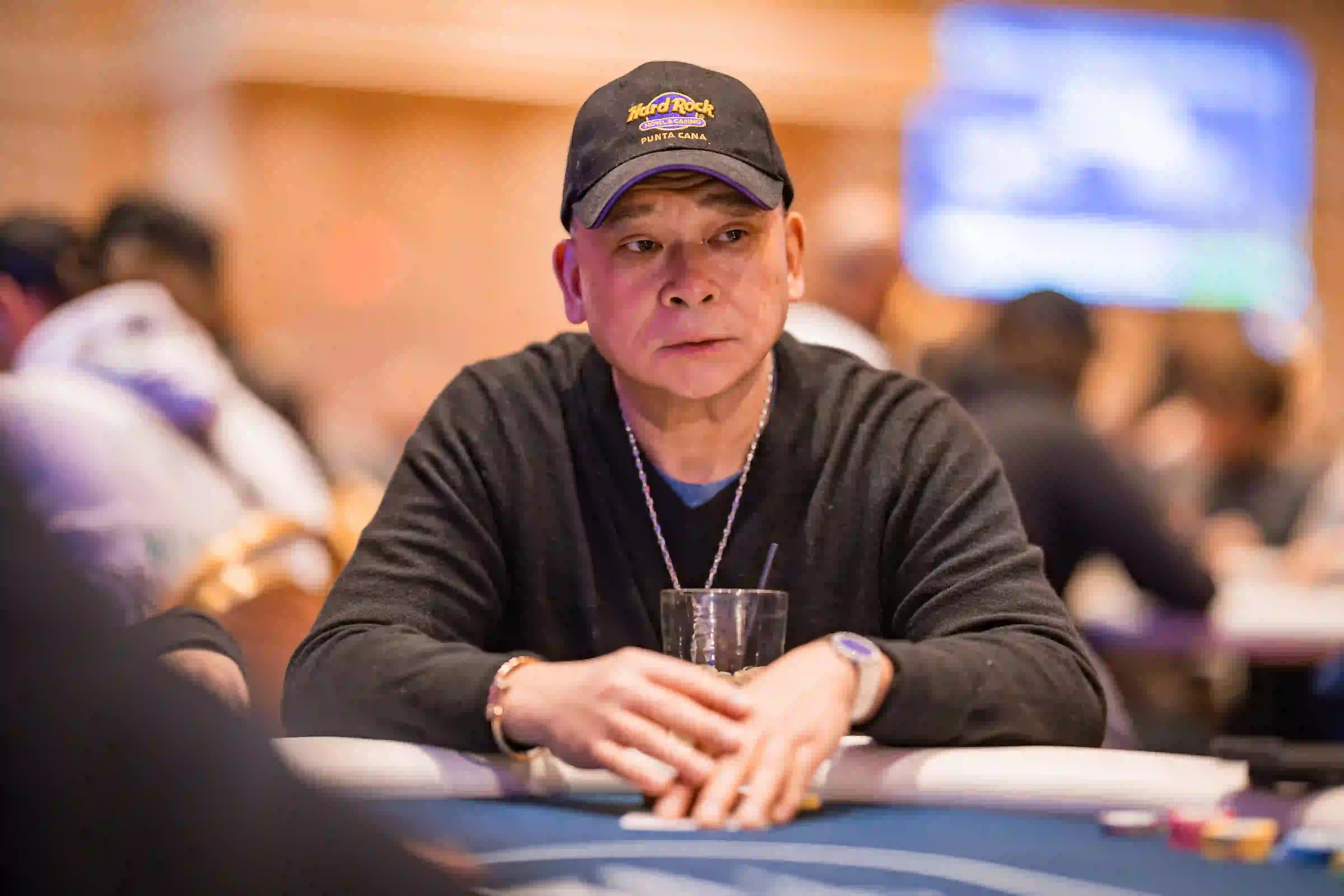 Johnny Chan wearing a Hard Rock Punta Cana cap and black sweater sits at the table with chips and a drink, focused on the action.