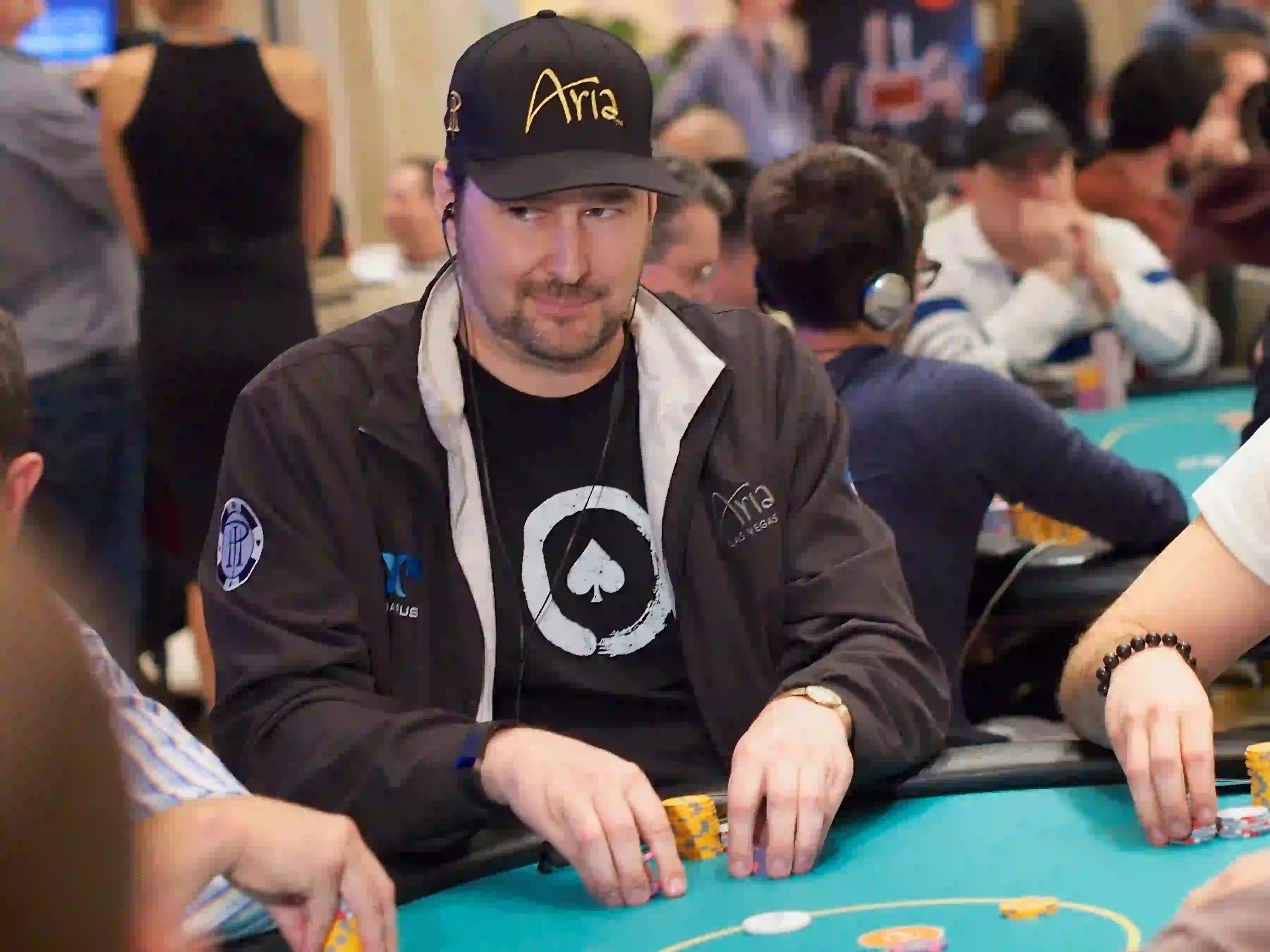 Phil Hellmuth sits at a poker table wearing an Aria cap and black jacket, holding chips during a live tournament