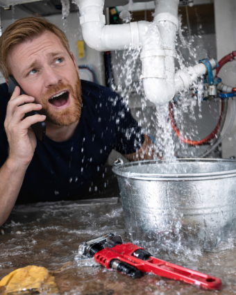 Plumber in uniform repairing pipes under a kitchen sink with tools and flashlight.