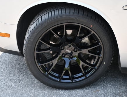 A close-up view of a black alloy wheel with a glossy finish, mounted on a white vehicle, showcasing the intricate spoke design and a tire with visible tread patterns.