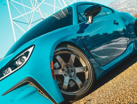 A close-up of a blue sports car with sleek aerodynamic design and large alloy wheels, set against a sunny outdoor backdrop with a tower structure.