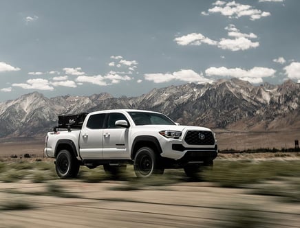 A white pickup truck driving past a mountainous landscape under a cloudy sky.