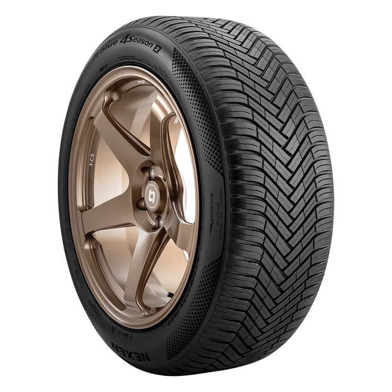 A close-up of a Nexen N'blue 4 Season 2 tire with a distinctive tread pattern, mounted on a bronze alloy wheel with a five-spoke design.