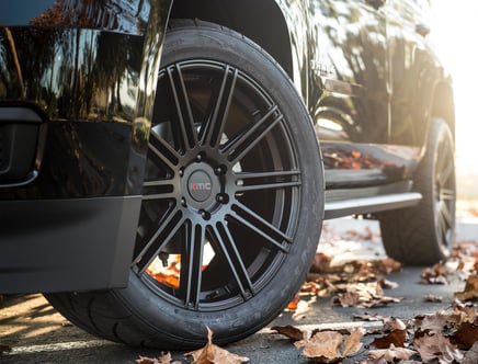Close-up of a car with a KMC wheel rim, surrounded by dry autumn leaves on the ground, reflecting the morning sunlight on a smooth, black surface.