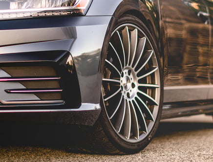 Close-up of a car's front wheel and fender, featuring a sleek alloy wheel design and a partial view of the headlight and bumper, set on a paved surface.