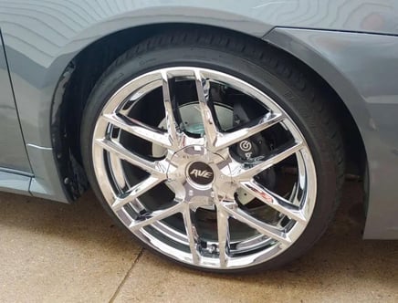 A chrome rim with intricate design on a car's wheel, featuring an AWE logo and visible brake caliper behind it.