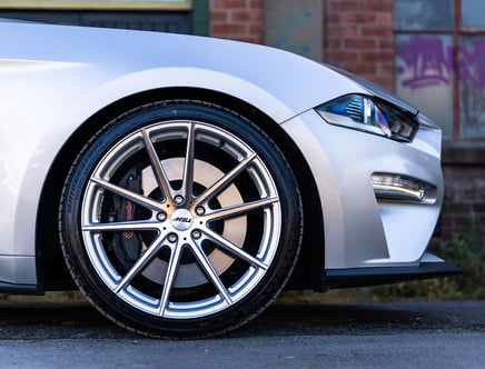 Close-up of a silver car's front wheel featuring a multi-spoke alloy rim and Brembo brake calipers, with part of the headlight and bumper visible, set against an urban background.