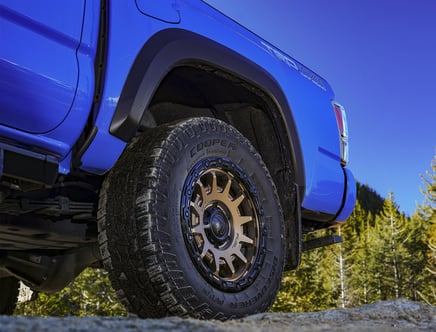 A close-up view of a blue truck's rear tire and wheel with a bold design, set against a clear blue sky and forest background.