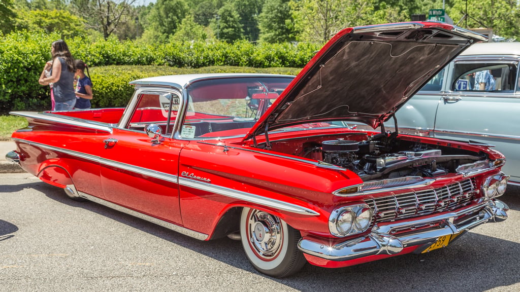 A classic red Chevrolet El Camino with its hood open, showcasing the engine and tucked wheel fitment, on display at a car show with trees and spectators in the background.