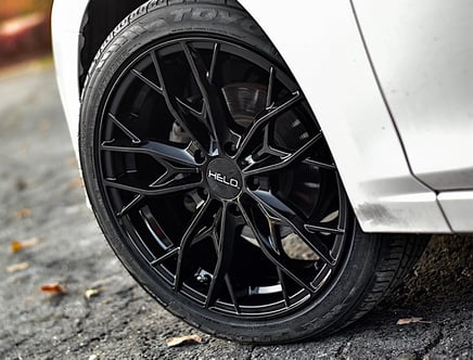 Close-up of a black Helo alloy wheel on a white car, parked on an asphalt surface with some leaves scattered around.