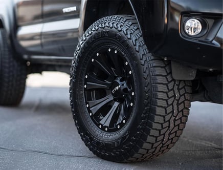 Close-up view of a black off-road vehicle tire with deep tread pattern and black alloy rim, parked on a gray surface.