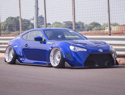 A modified blue sports car with large rims and lowered suspension parked on a racetrack with a metal fence in the background.