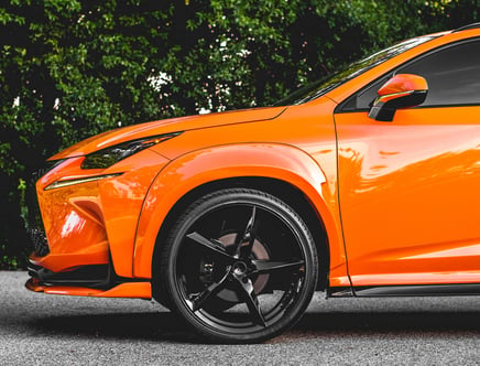Close-up of an orange SUV with black rims and tinted windows, parked on a paved area with greenery in the background.