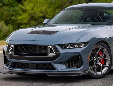 A blue sports car with a sleek, angular front design, featuring distinctive LED headlights and a prominent grille. The car has red brake calipers visible behind alloy wheels. Trees are in the background.