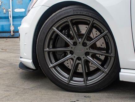 Close-up of a car's wheel featuring a grey multi-spoke alloy rim with a TSW logo, paired with a tire marked "Azenis," against a blue industrial backdrop.