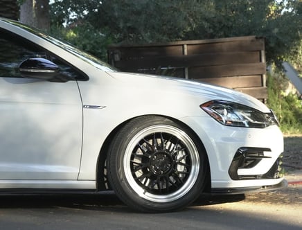 Front side view of a white car with black rims and a sleek design, parked on a street next to wooden fencing and greenery.