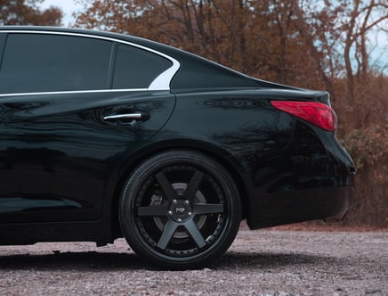 Rear view of a black car with tinted windows, featuring a bright red taillight and black alloy wheels, parked on a gravel surface with autumn foliage in the background.
