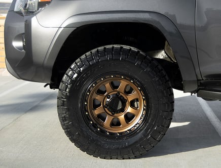 Close-up of a car's front wheel featuring a gold alloy rim and a thick off-road tire, set against a gray car body in a parking lot.