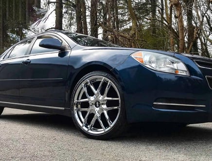 A blue car with shiny chrome rims parked on a road, surrounded by trees in the background.