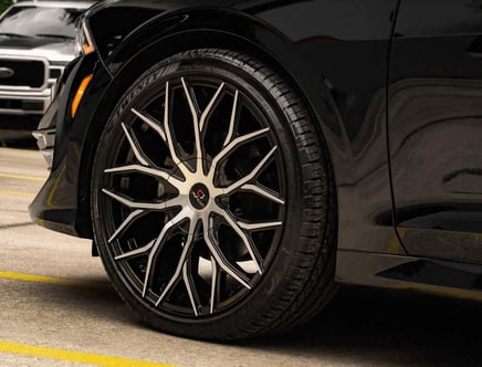 Close-up of a black car's wheel with intricate, angular alloy rim design, parked on a concrete surface with yellow lines.