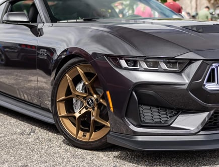 Close-up of the front end of a gray sporty car with distinctive angular headlights and bronze alloy wheels, parked on a road surface.