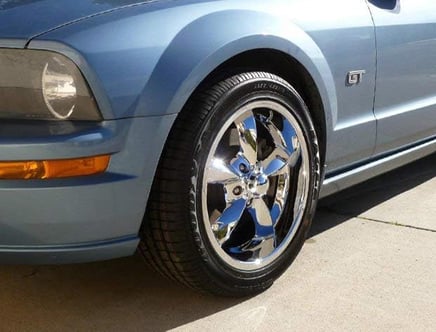 Close-up of a blue car's front left wheel with shiny chrome rims, parked on a concrete surface, showing the GT badge on the car's side.
