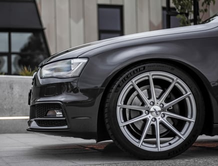 Close-up view of the front left side of a dark-colored car featuring a sleek headlight and a prominently displayed alloy wheel with Michelin tires, set against an urban background.