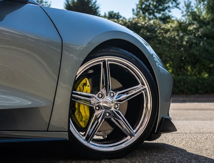 Close-up of a gray sports car's wheel featuring shiny chrome spokes and a yellow brake caliper, set against a background of trees and blue sky.