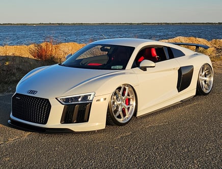 A white sports car parked near a waterfront with a clear sky in the background, featuring red interior seats and sleek design elements.