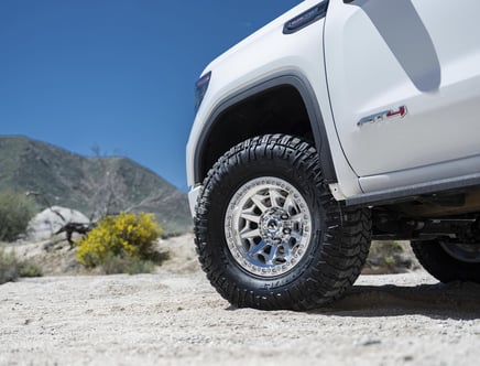 Close-up of a white off-road vehicle's tire on sandy terrain, with mountainous landscape in the background under a clear blue sky.