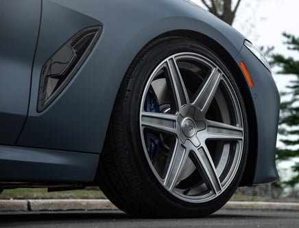 Close-up of a gray car's wheel and tire with a sleek alloy rim, featuring a side vent on the car's body adjacent to the wheel.