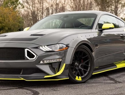 A customized gray sports car with neon yellow accents on the side mirror, wheels, and front bumper, parked on a paved surface with trees in the background.