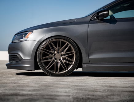 Close-up view of the front side of a grey car with large multi-spoke alloy wheels, parked on a concrete surface under a clear blue sky.