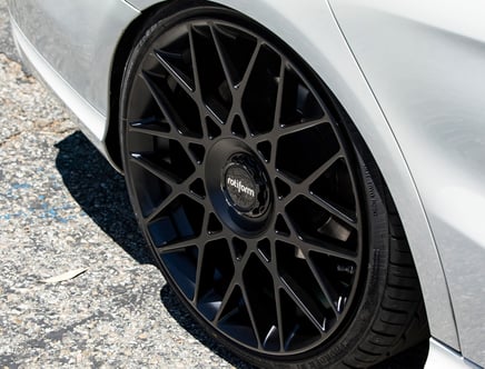 Close-up of a black, multi-spoke car wheel on a gray vehicle, parked on a textured surface.