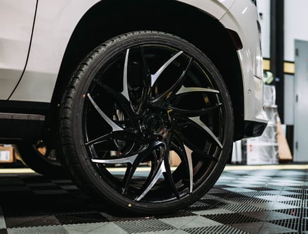 Close-up of a black alloy wheel with intricate design on a white vehicle, parked on a checkered floor in a garage setting.