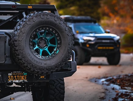 Close-up of a rugged off-road tire and wheel on a vehicle, with another off-road vehicle blurred in the background.